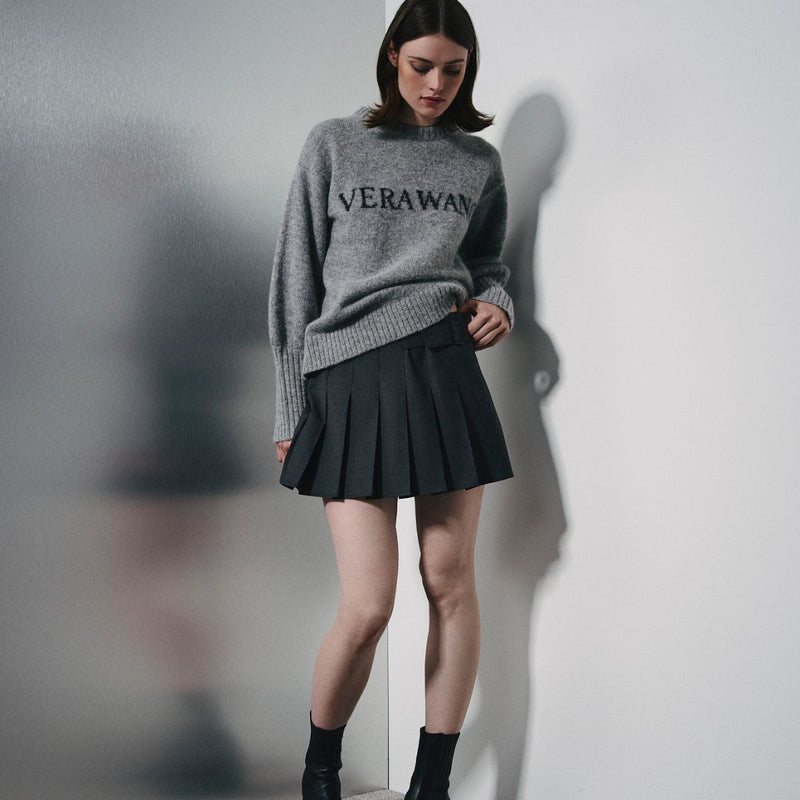 A model stands against a white wall wearing a gray knit sweater with 'VERAWAN' printed across the chest and a black pleated mini skirt.