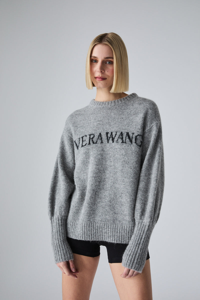 A model with blonde bob-cut hair wearing a heather gray knit sweater with 'VERAWANG' printed in black across the chest, paired with black shorts, against a solid gray studio background.