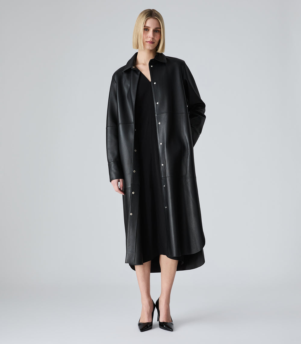 A model wearing a long black leather-like button-front coat over a black dress, standing against a light gray studio background.