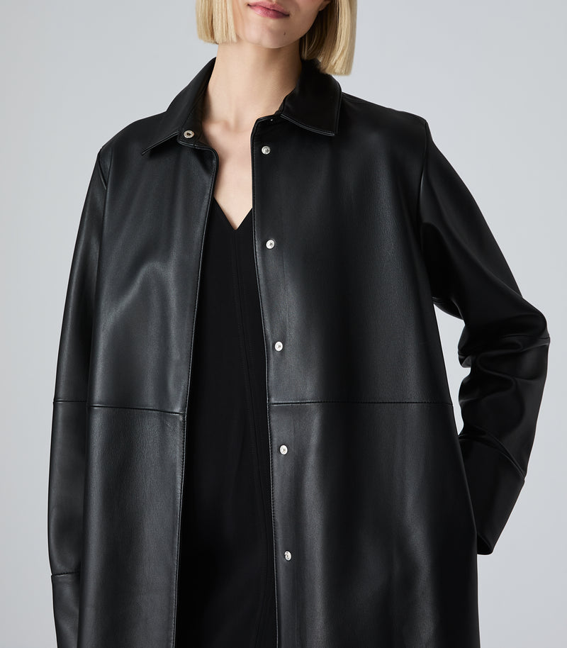 A model wearing an oversized black faux-leather button-up shirt jacket over a black top, shown from the chin down against a light gray studio background.