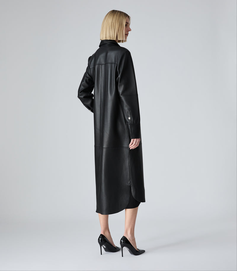 A woman with short blonde hair is shown from the back wearing a long black leather coat and black high heels against a light gray studio background.