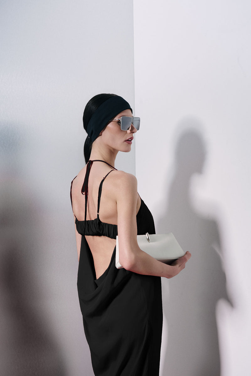 A woman in a black strappy dress with an open back, wearing a black headscarf and silver-framed sunglasses, holding a white clutch bag against a white wall.