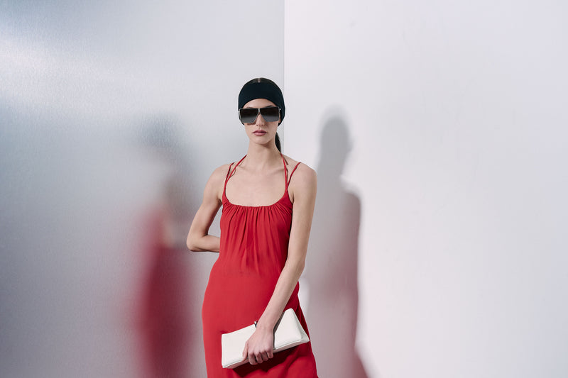 A woman wearing a red spaghetti-strap midi dress, black sunglasses, and a black head scarf holds a white clutch bag against a minimalist white wall with a soft shadow.