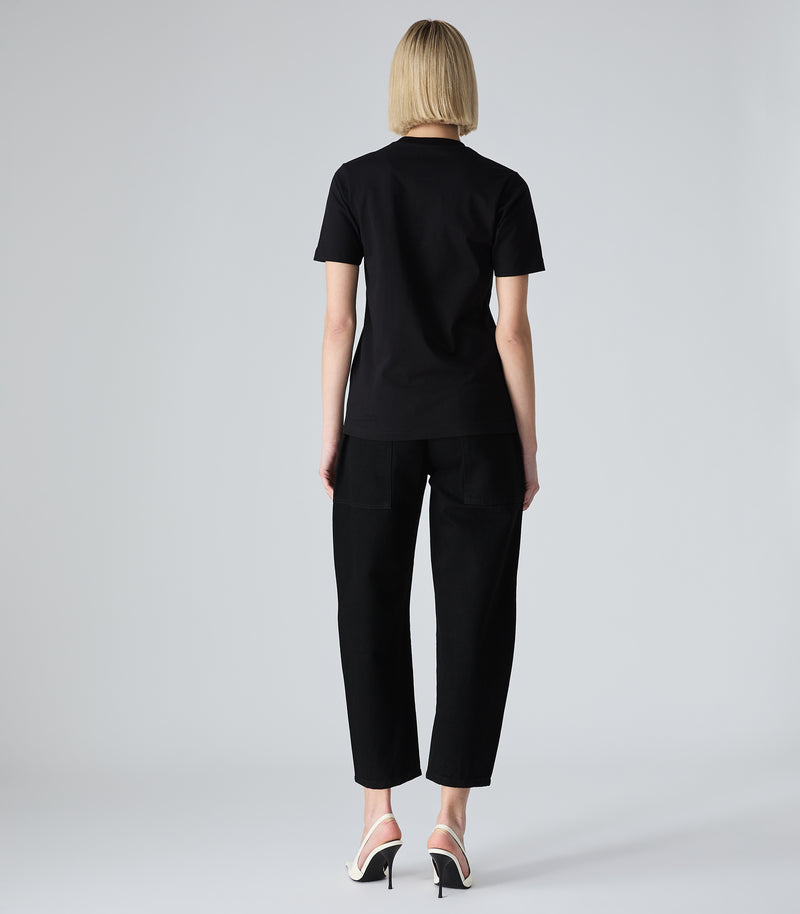Female model wearing a slim-fit black t-shirt, paired with black trousers and white heels, against a white background.