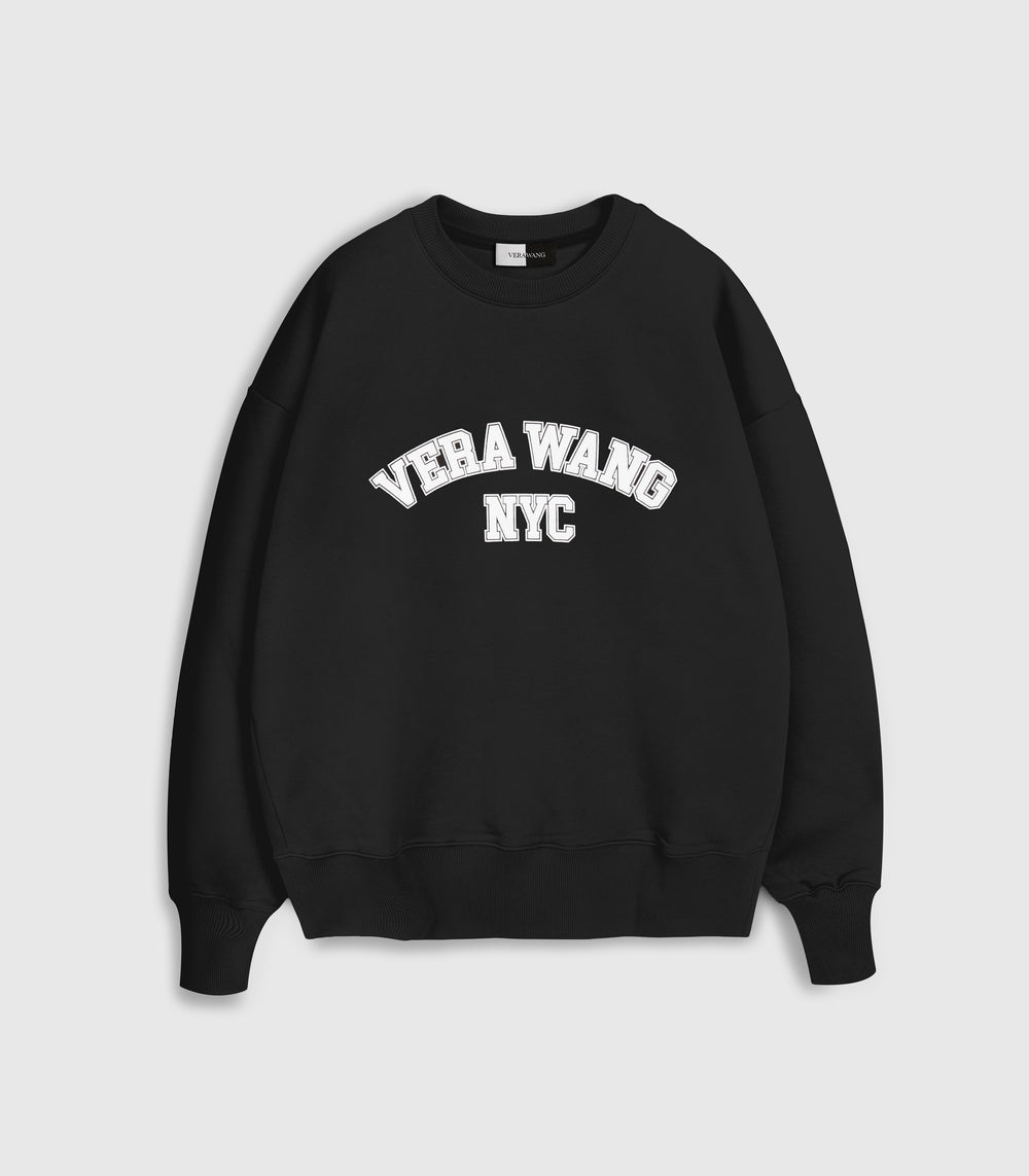Flat lay product view of a relaxed-fit black crewneck sweatshirt featuring an arched varsity-style 'VERA WANG NYC' graphic printed in white block lettering with a subtle outline across the chest, complete with a ribbed collar, hem, and cuffs against a light grey background.