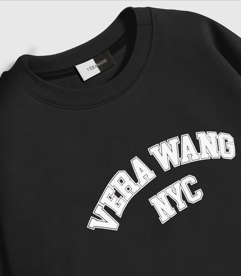 Close-up, angled product view of a black crewneck sweatshirt, showcasing the white printed 'VERA WANG NYC' varsity block graphic across the front, a chunky ribbed knit collar, and a split black-and-white inner neck brand label against a light grey background.