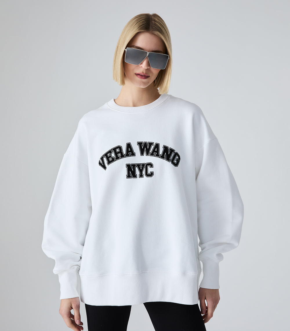 Half-length view of a model wearing an oversized white crewneck sweatshirt featuring 'VERA WANG NYC' in black collegiate arch lettering across the chest. The jumper is styled with black leggings and futuristic silver shield sunglasses, against a white background.