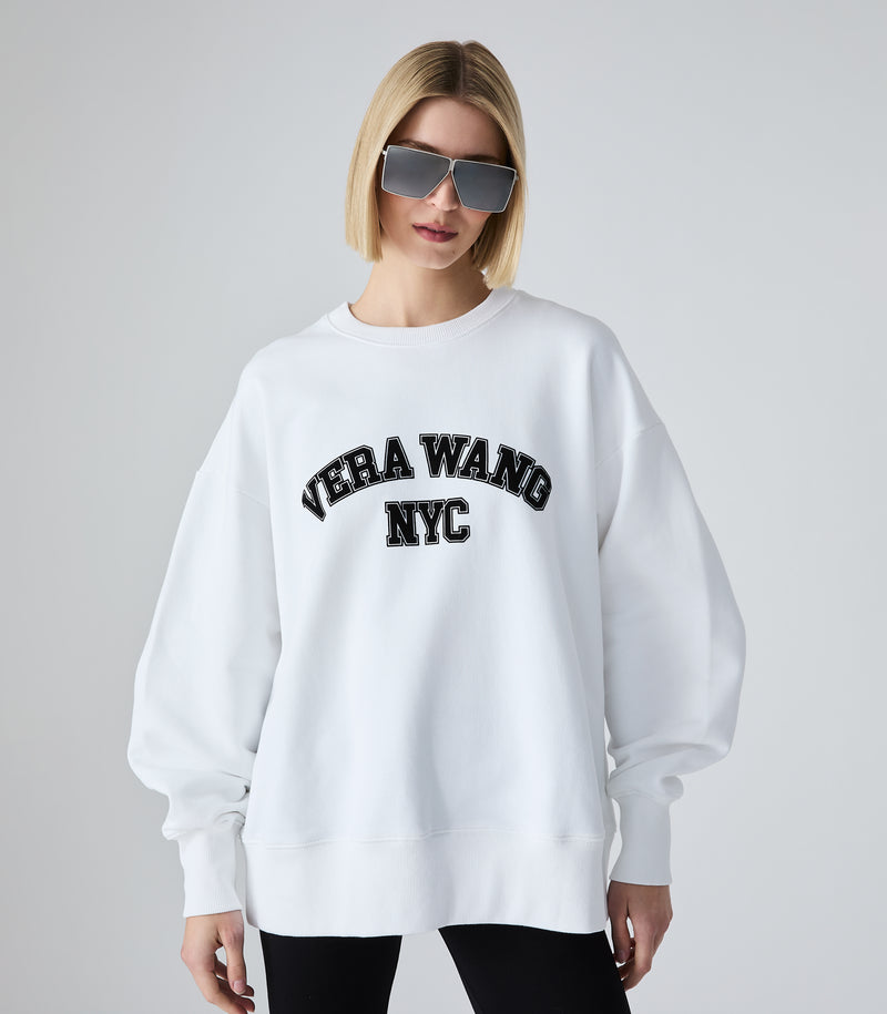 Half-length view of a model wearing an oversized white crewneck sweatshirt featuring 'VERA WANG NYC' in black collegiate arch lettering across the chest. The jumper is styled with black leggings and futuristic silver shield sunglasses, against a white background.