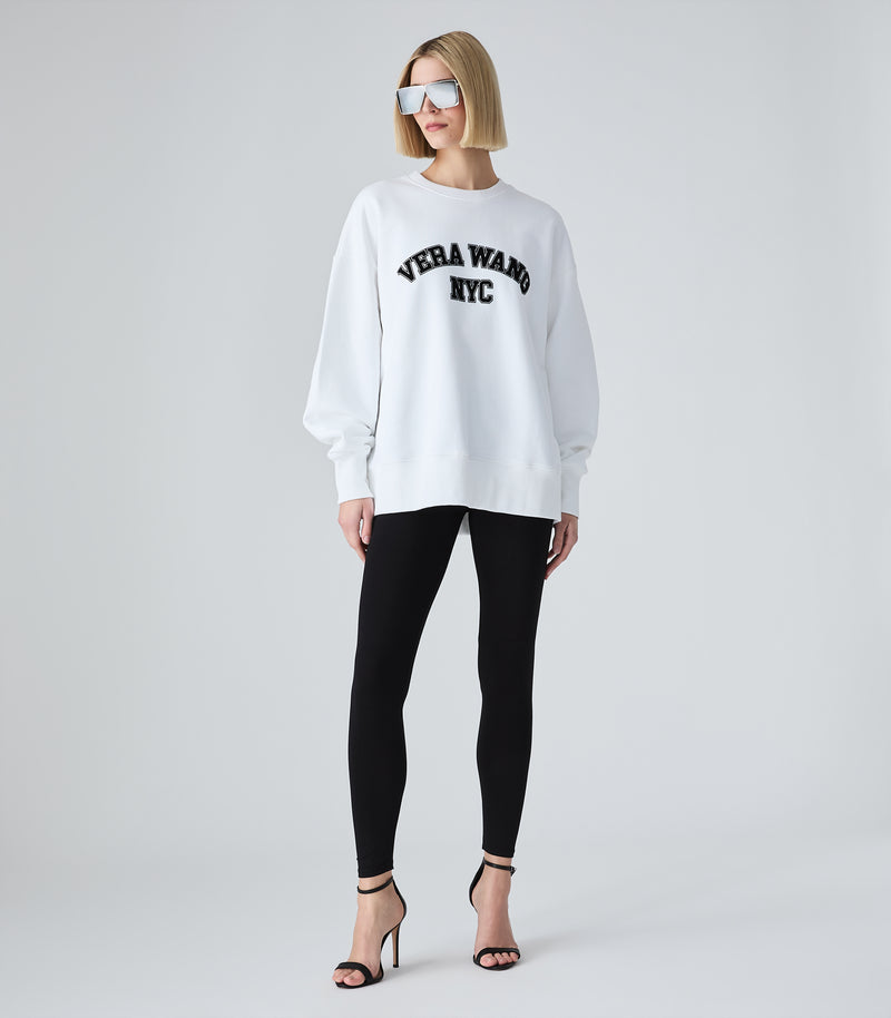 Full-length view of a model wearing an oversized white crewneck sweatshirt featuring 'VERA WANG NYC' in black collegiate arch lettering across the chest. The jumper is styled with black leggings, futuristic silver shield sunglasses, and black strappy stiletto heels against a white background.
