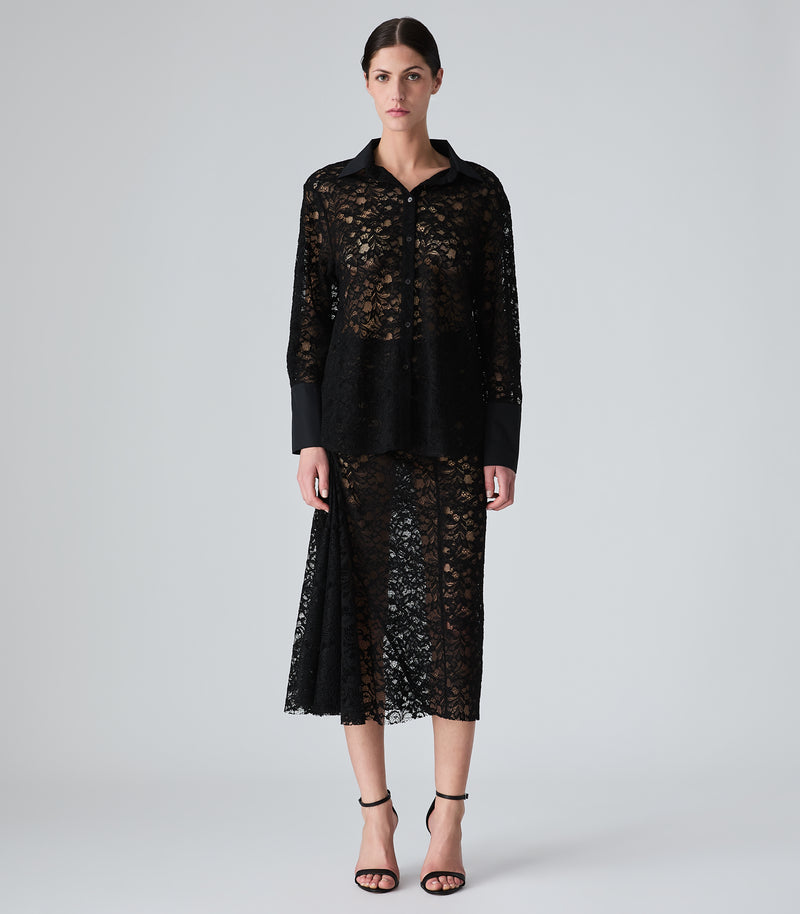 A model stands against a light gray studio background wearing a black sheer lace three-piece set consisting of a long-sleeve collared top, a matching peplum-style overlay, and a midi skirt, paired with black strappy heels.