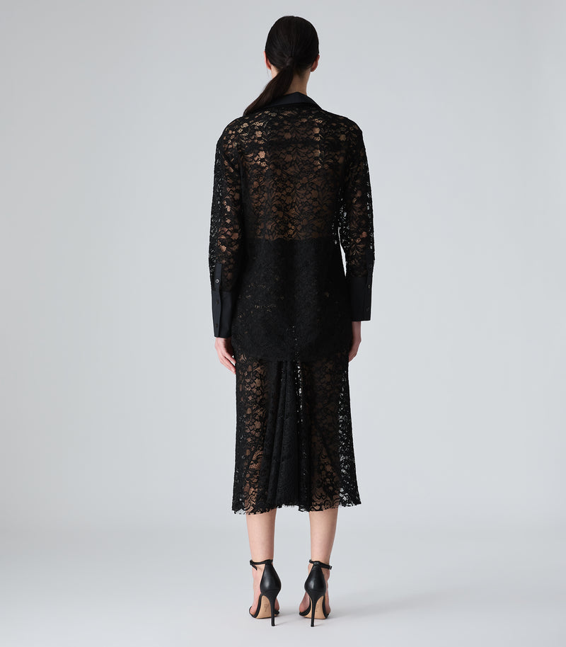 A model viewed from the back wearing a black lace long-sleeve blazer and matching midi skirt, paired with black high-heeled sandals, against a light gray studio background.