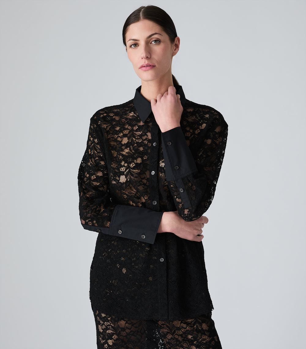 A woman modeling a black long-sleeve lace button-up shirt with solid black collar, cuffs, and button placket, photographed against a light gray studio background.