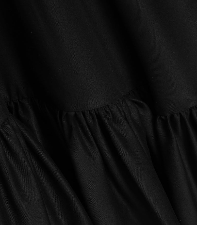 Macro close-up view of the skirt on a black smock dress, highlighting the fine cotton-blend fabric texture and smooth finish. The shot focuses on a dense row of delicate gathers running along a horizontal seam that transitions into a voluminous ruffled tier.