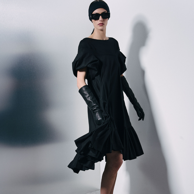 A model wearing a black off-the-shoulder midi dress with dramatic puffed sleeves and a layered hem, styled with matching long black gloves, a black beanie, and black sunglasses against a white wall with a strong shadow.