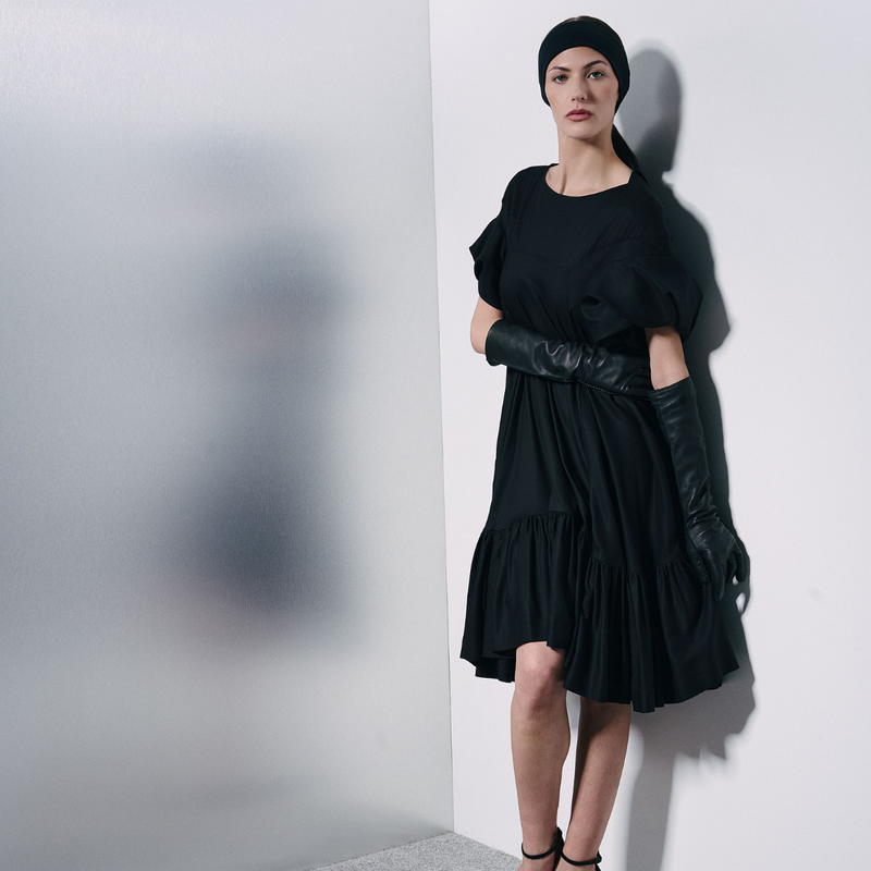 A model wearing a black tiered midi dress with short puff sleeves, black elbow-length gloves, and a black beanie, posing against a white wall.