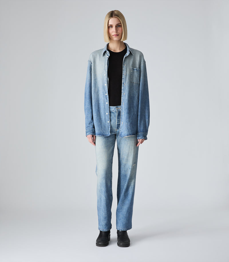 A model stands against a light gray studio background wearing a light-wash blue denim button-up shirt over a black top, matching light-wash blue jeans, and black boots.