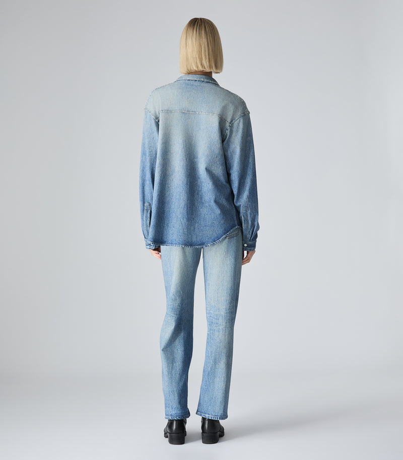 A model viewed from behind wearing a light blue denim long-sleeve button-up shirt and matching light blue denim jeans, paired with black shoes, against a light gray studio background.