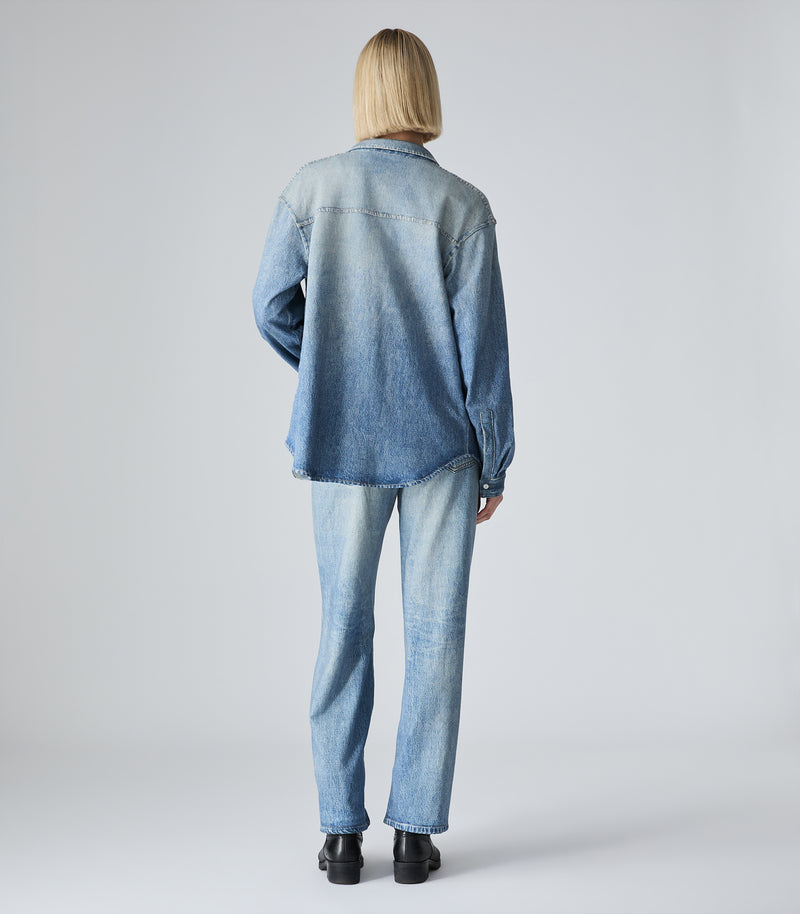 A model viewed from behind wearing a light blue denim button-up shirt and matching light blue denim jeans, paired with black shoes, against a light gray studio background.