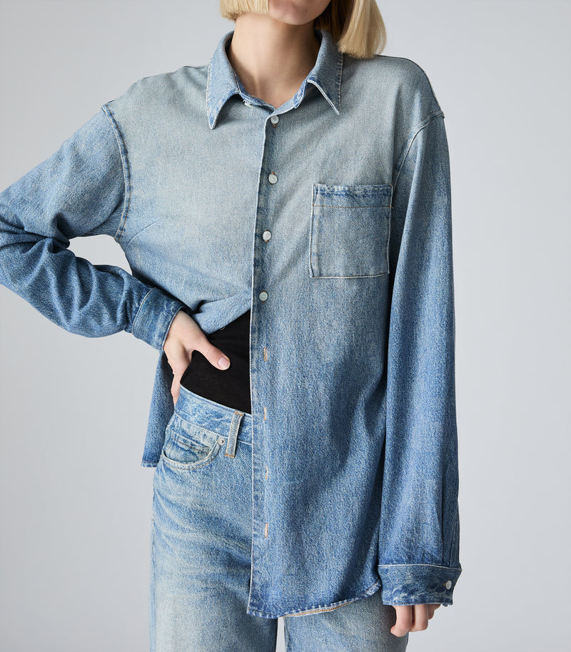 A model wearing an oversized light-wash blue denim shirt with a chest pocket, styled over a black top and paired with matching denim jeans against a plain light gray background.
