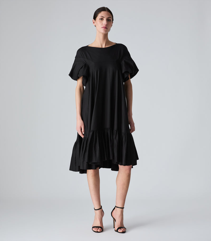 Full-length view of a model wearing a knee-length black smock dress featuring a relaxed, fluid silhouette, a high boat neckline, and dramatic voluminous ruffled short sleeves. The dress is designed with a tiered, heavily gathered ruffle hemline and is styled with black ankle-strap heels against a light grey background.