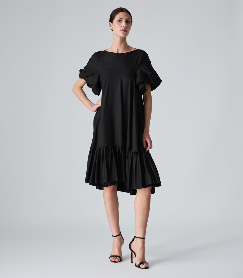 Full-length view of a model wearing a knee-length black smock dress featuring a relaxed, fluid silhouette, a high boat neckline, and dramatic voluminous ruffled short sleeves. The dress is designed with a tiered, heavily gathered ruffle hemline and is styled with black ankle-strap heels against a light grey background.