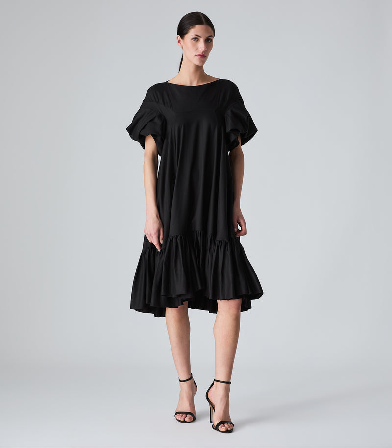 Full-length view of a model wearing a knee-length black smock dress featuring a relaxed, fluid silhouette, a high boat neckline, and dramatic voluminous ruffled short sleeves. The dress is designed with a tiered, heavily gathered ruffle hemline and is styled with black ankle-strap heels against a light grey background.