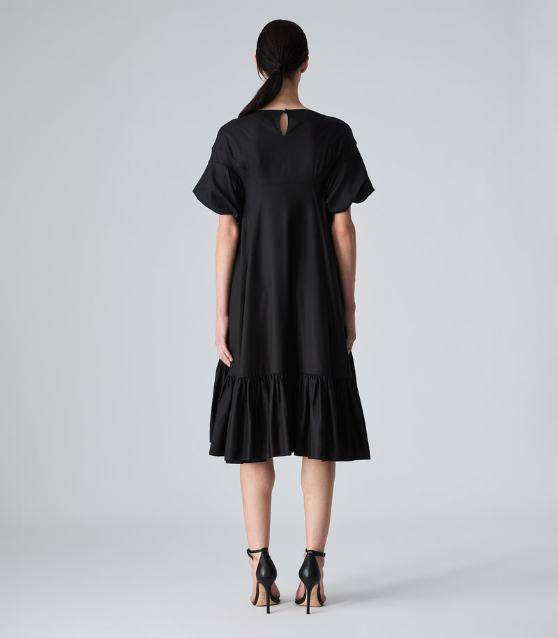 Full-length rear view of a model wearing a knee-length black smock dress featuring a loose, fluid silhouette, a high back neckline with a delicate keyhole cutout closure, and voluminous ruffled short sleeves. The dress is designed with a gathered, tiered ruffle hemline and is styled with black ankle-strap heels against a light grey background.