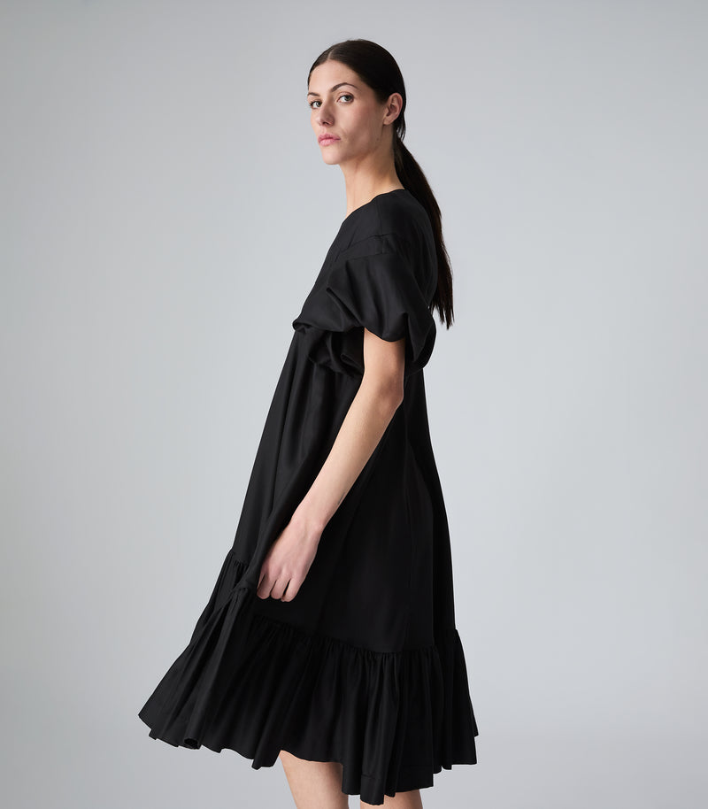 Full-length side-profile view of a model wearing a knee-length black smock dress featuring a relaxed, tent-style silhouette, dramatic voluminous ruffled short sleeves, and a tiered ruffled hemline that shows fluid movement. The model is captured mid-turn looking over her shoulder, presenting the garment against a light grey background.
