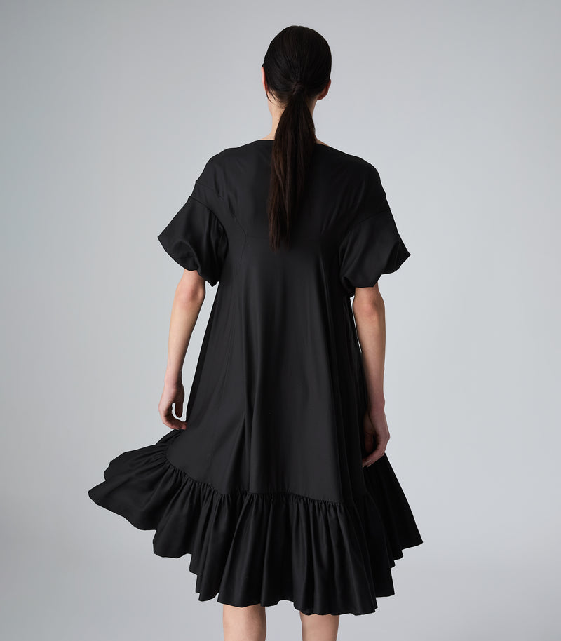 Three-quarter rear view of a model wearing a knee-length black smock dress, showcasing the fluid movement of the garment. The dress features a relaxed, voluminous silhouette with contoured back panel seams, dramatic puffed short sleeves, and a tiered, heavily gathered ruffle hemline that flares out dynamically. The model is presented from the back against a light grey background.