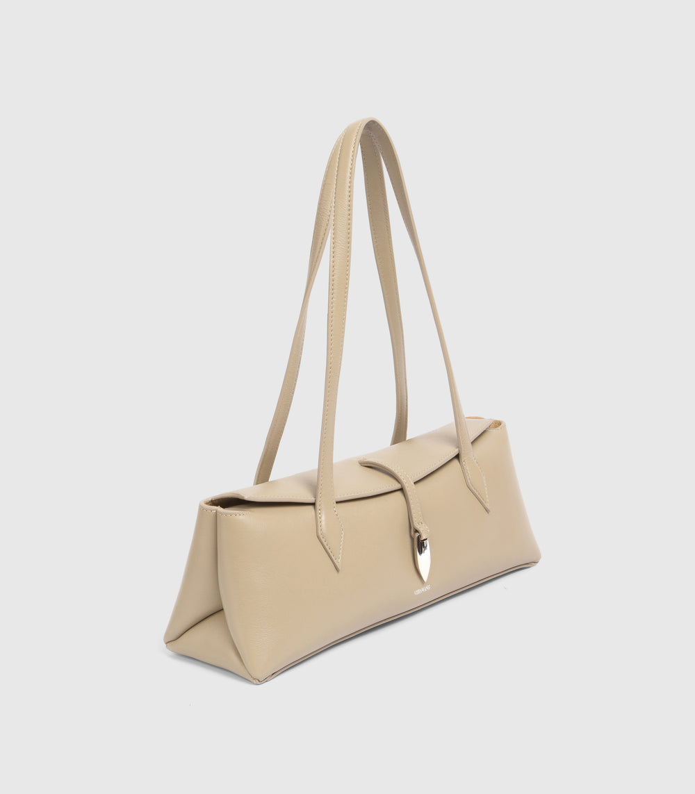 A structured beige leather shoulder bag with long straps and a small metallic shield charm, shown on a light gray background.