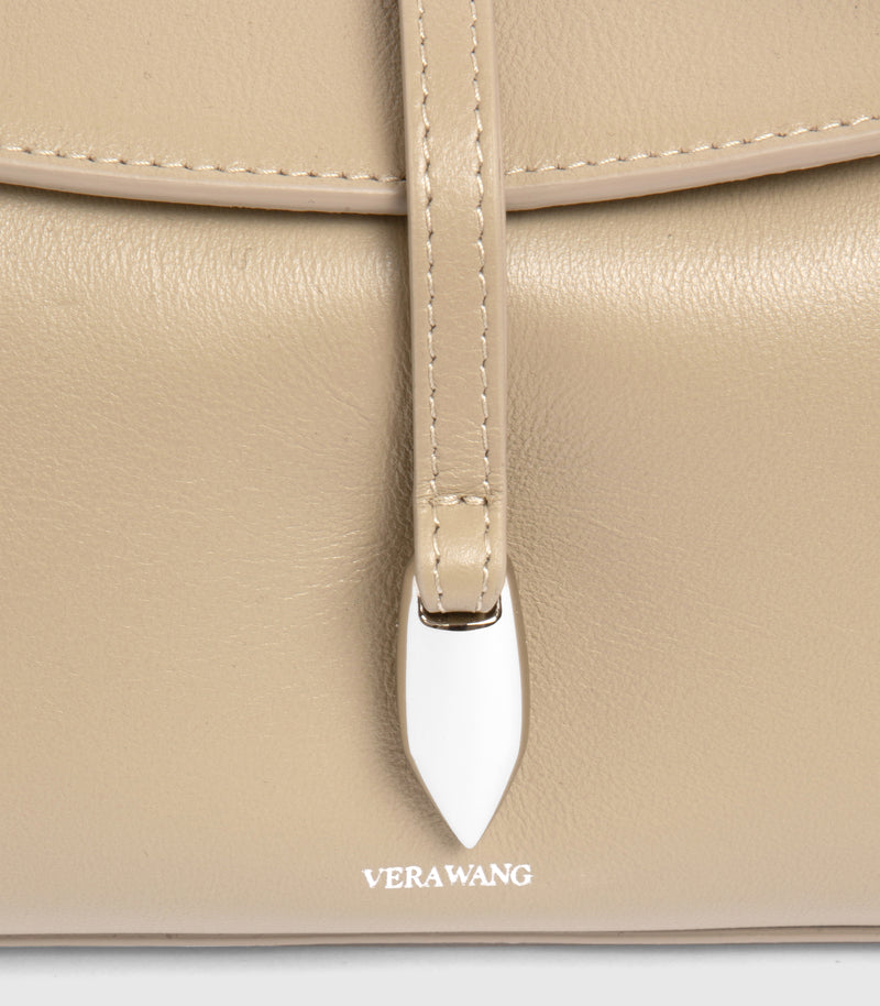Close-up of a beige leather handbag with a vertical strap and a white teardrop-shaped metal accent, featuring the VERA WANG logo at the bottom.