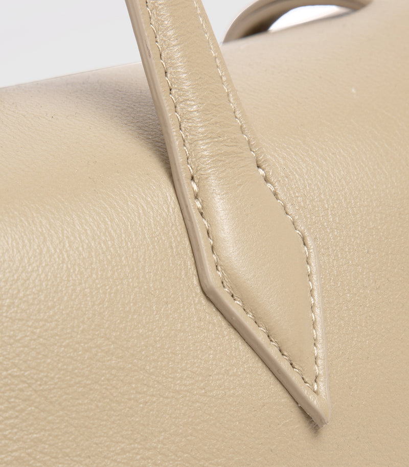 Close-up of a light beige pebbled leather handbag showing a stitched leather strap attachment and part of a top handle.