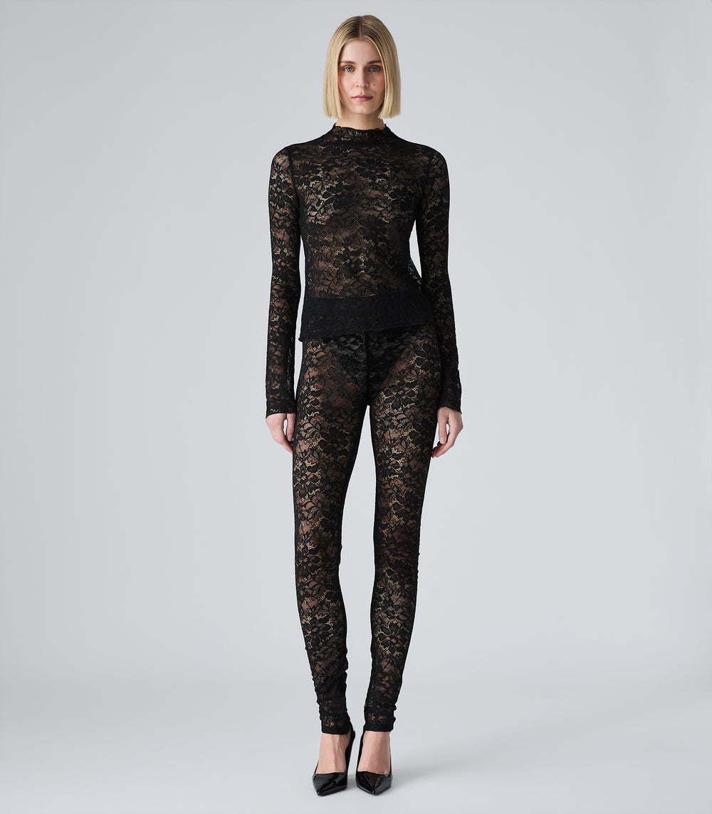 Full-length studio view of a model showcasing a pair of sheer black lace leggings. The focus is on the skinny-fit, high-rise design crafted from intricate floral lace, displaying a semi-translucent overlay style. The leggings are layered over dark undergarments and pool slightly at the ankles, styled as a co-ord set with a matching long-sleeved lace top and black patent stiletto pumps against a seamless light grey background.