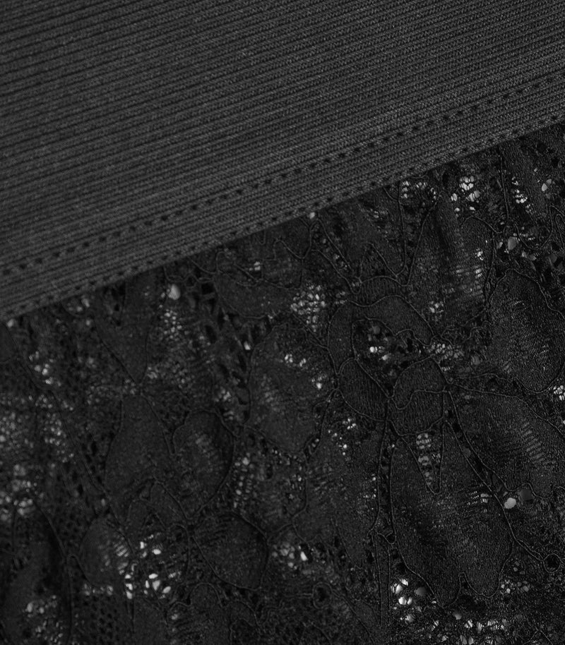 An extreme close-up product view of a pair of black leggings, showcasing the construction of the waistband. The macro shot highlights a wide, solid black elastic band with a finely ribbed texture and clean double-needle topstitching where it securely joins the sheer, gathered black floral lace body fabric.