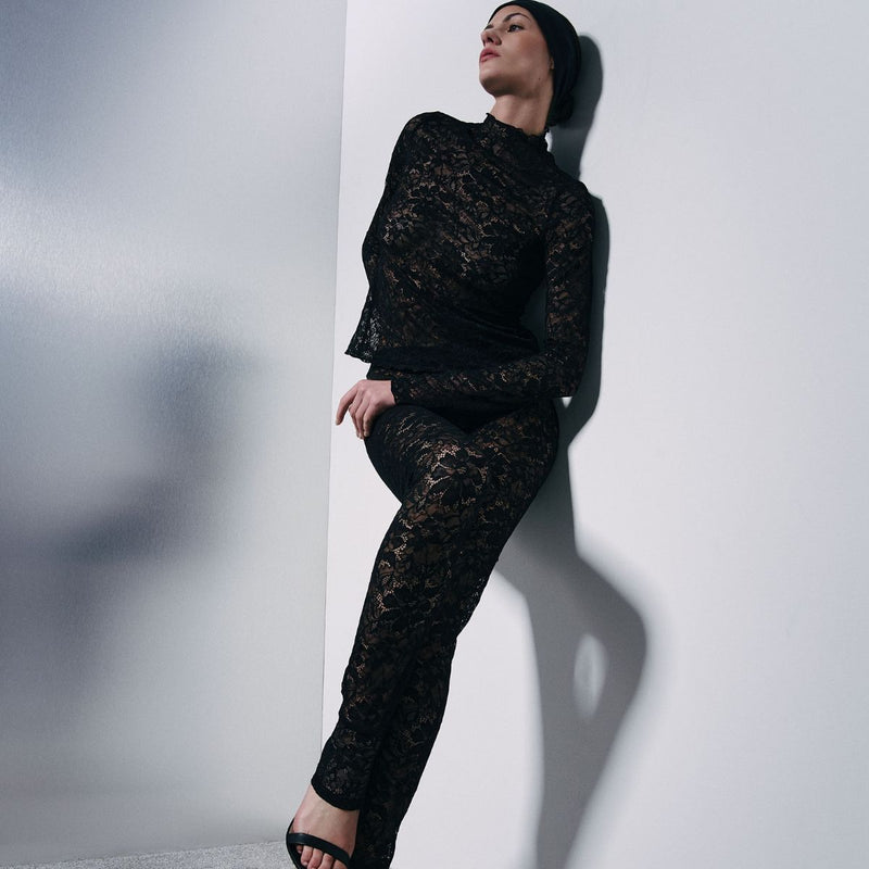 A model wearing a black lace long-sleeve top and matching black lace pants, posing against a white wall.