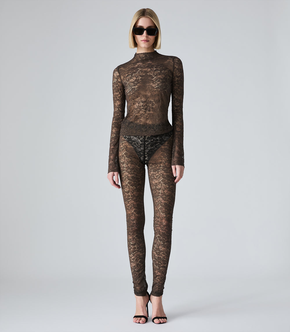 A model wearing a sheer brown lace long-sleeve mock-neck top and matching lace leggings, styled with black sunglasses and black high-heeled sandals against a light gray studio background.