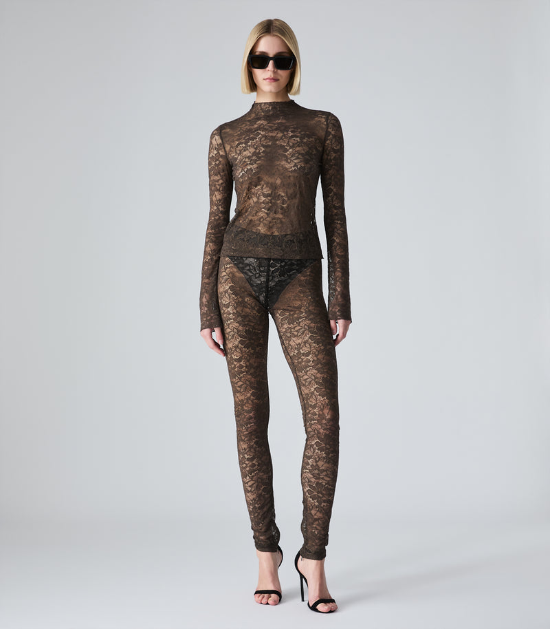 A model wearing a sheer brown lace mock-neck long-sleeve top and matching lace pants, styled with black sunglasses and black high-heeled sandals against a light gray studio background.