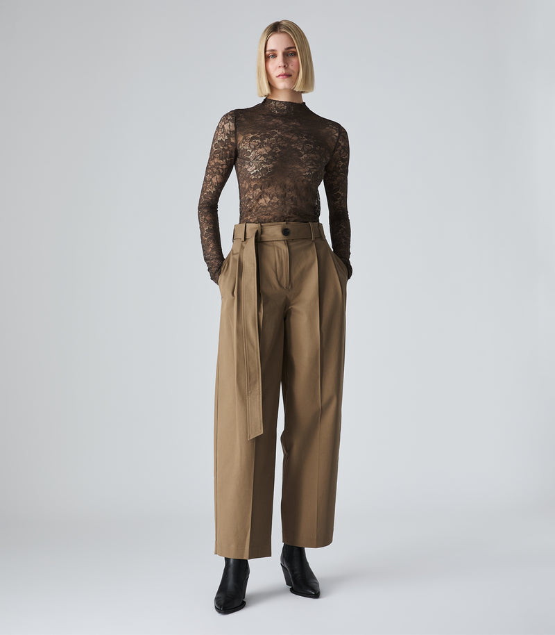 A model stands against a light gray studio background wearing a sheer black lace long-sleeve mock-neck top and high-waisted tan wide-leg trousers with a matching fabric tie belt, paired with black ankle boots.