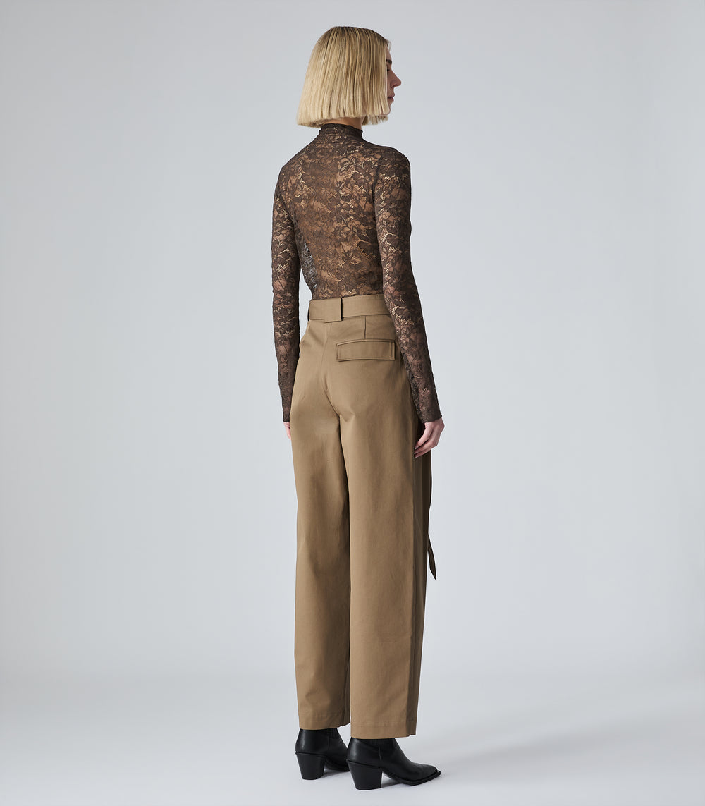 A model shown from the back wearing a sheer brown lace long-sleeve mock neck top and high-waisted tan wide-leg trousers, paired with black ankle boots, against a light gray studio background.