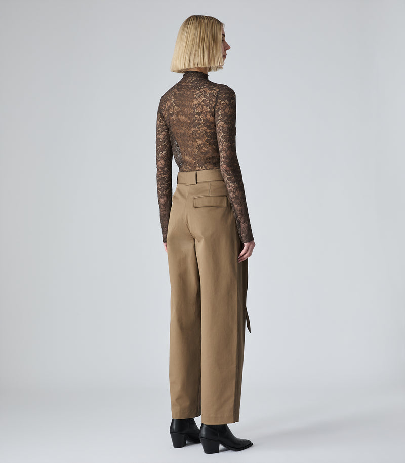 A model shown from the back wearing a sheer brown lace long-sleeve mock neck top and high-waisted tan wide-leg trousers, paired with black ankle boots, against a light gray studio background.