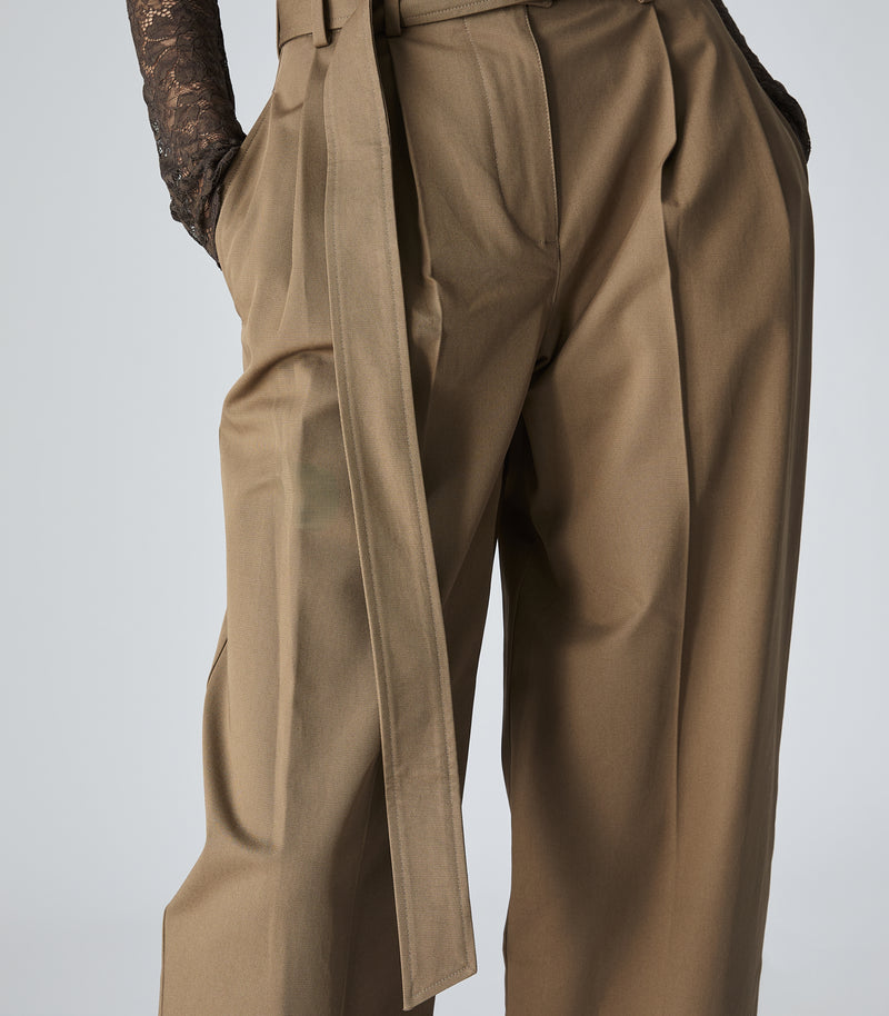 Close-up of a model wearing high-waisted, wide-leg trousers in a warm khaki-brown color. The pants feature a matching fabric belt with a long wrap-around sash, pleated front, and side pockets.