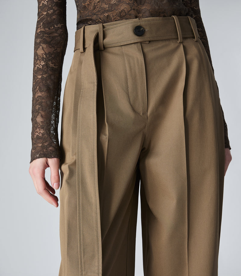 A close-up of a model wearing high-waisted, wide-leg trousers in a warm khaki-brown color, styled with a matching waist belt and a sheer lace long-sleeve top against a light gray studio background.