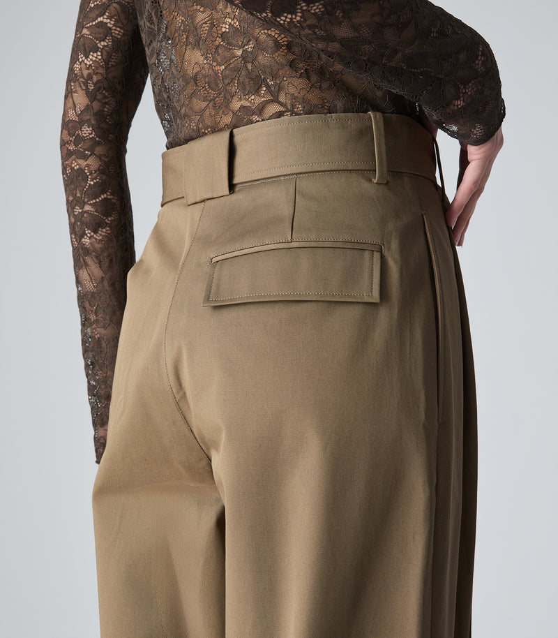 A model shown from the back wearing a sheer brown lace long-sleeve mock neck top and high-waisted tan wide-leg trousers, against a light gray studio background.