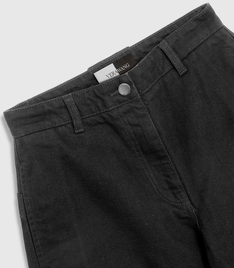 Close-up, angled product view of the waistband on a pair of washed black denim jeans, showcasing a metallic silver button hardware closure, belt loops, tonal stitching, and a woven black-and-white 'VERA WANG' inner waist label against a light grey background.