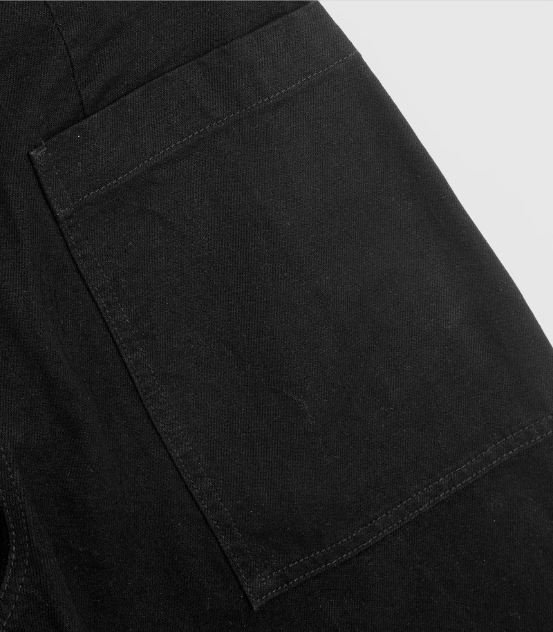 Close-up product view of a pair of black denim barrel jeans, highlighting the structured lines of a large square patch pocket, tonal topstitching, and the woven diagonal texture of the premium cotton twill denim fabric.