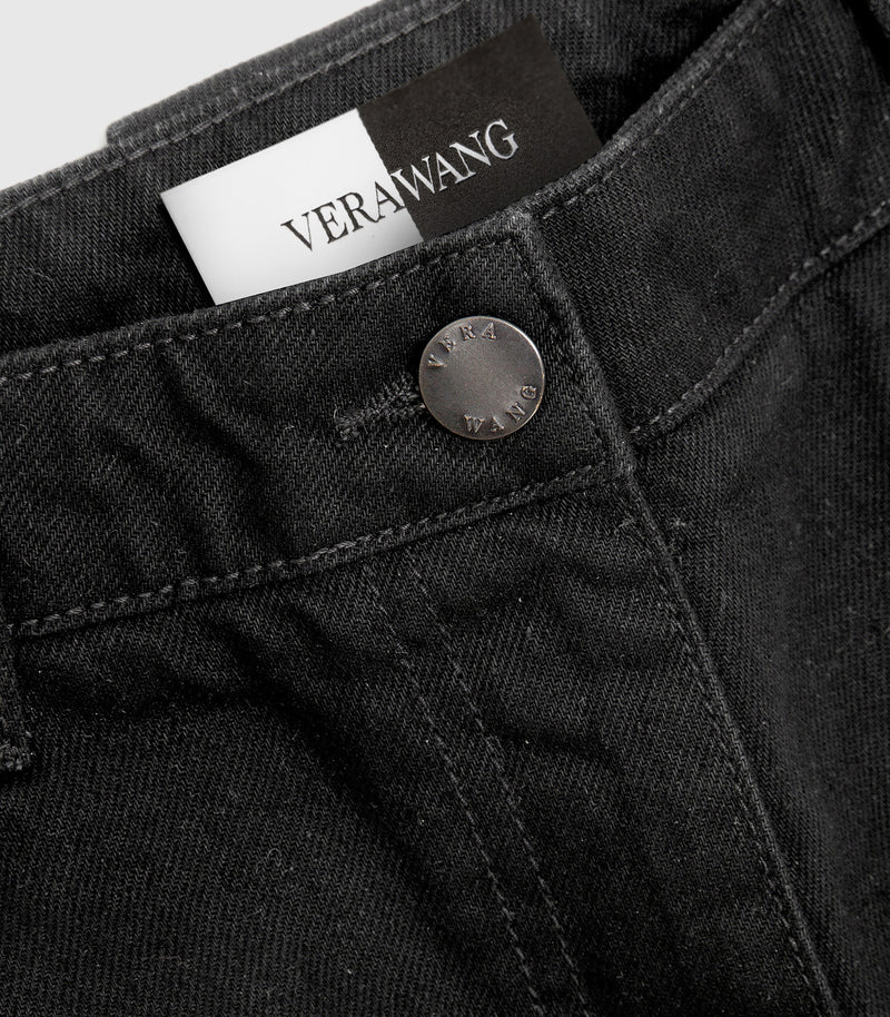 Extreme close-up product view of the front waistband on a pair of washed black denim jeans, showcasing a metallic button engraved with 'VERA WANG' typography, a detailed look at the buttonhole stitching, and a peek of the woven black-and-white inner tag against a light grey background.