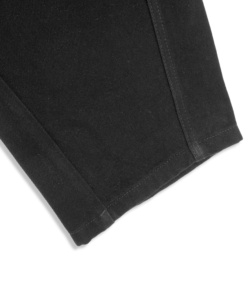 Close-up product view of the cropped leg hem on a pair of black denim jeans, highlighting the clean folded hemline stitch, structured curved side seams, and the diagonal weave of the premium cotton twill denim fabric against a light grey background.