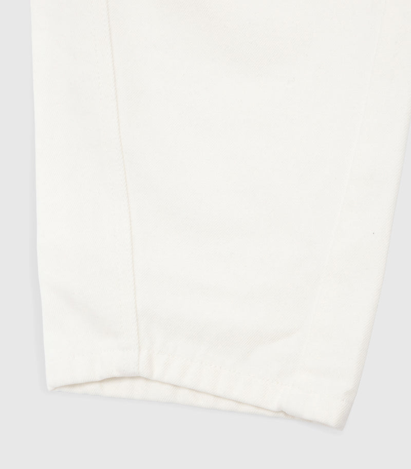 Close-up product view of the hem and curved leg seam on the ecru jeans, showcasing the textured cotton twill denim fabric and tonal stitching against a white background.