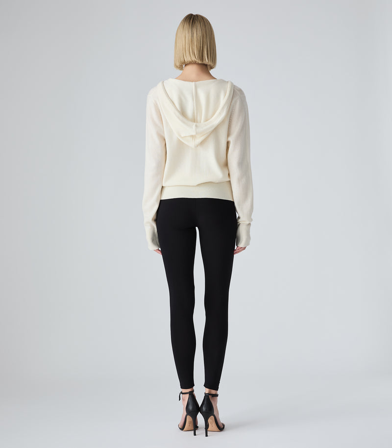 A woman viewed from behind wearing a cream-colored hooded sweatshirt, black leggings, and black high-heeled sandals against a light gray studio background.