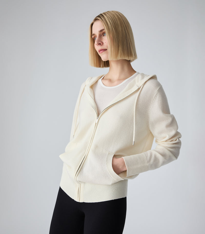 A woman with a short blonde bob wearing a cream-colored zip-up hoodie over a white t-shirt and black pants, photographed against a neutral light gray background.